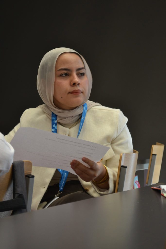 A woman wearing a white hijab and blazer sits at a table, holding a sheet of paper and looking to the side. She wears a blue lanyard and is in a conference or meeting room.
