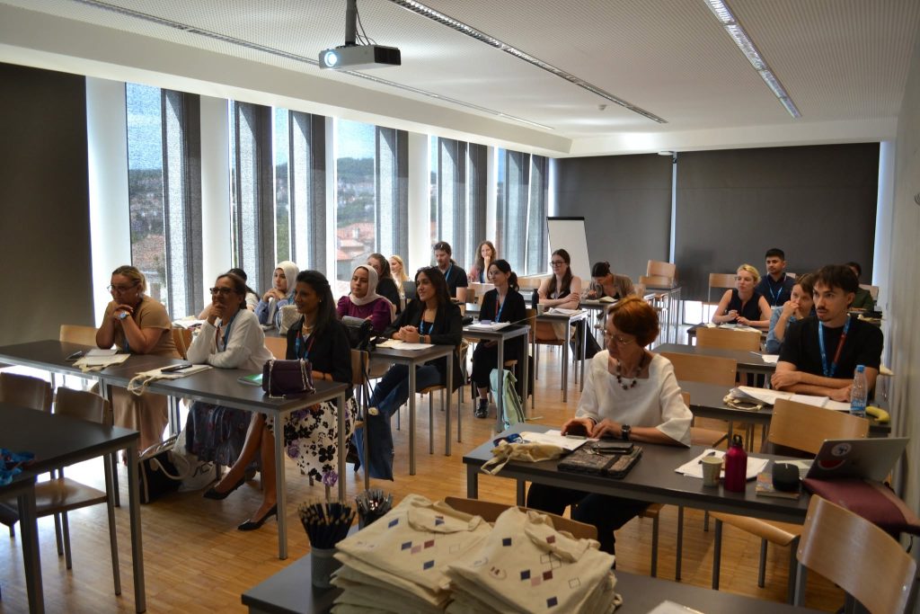 A diverse group of adults sit at desks in a classroom, attentively listening to a presentation. Sunlight streams through tall windows, and notebooks and bags are on the desks.