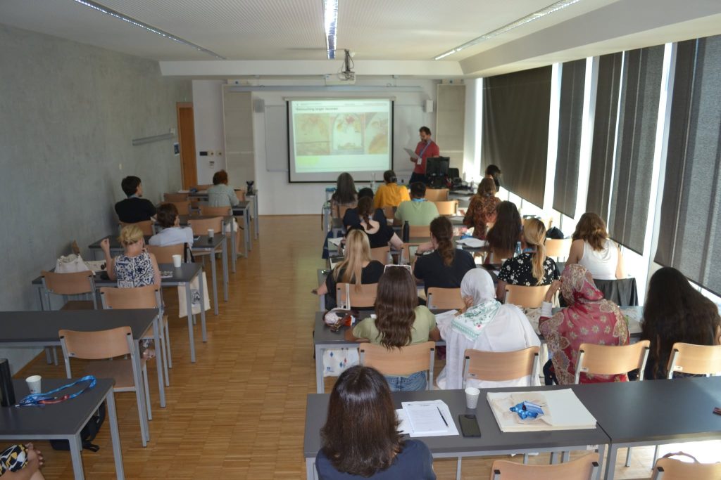A classroom with students seated at desks facing a teacher giving a presentation at the front. A projector displays images on a screen. The room has large windows with blinds and a wooden floor.