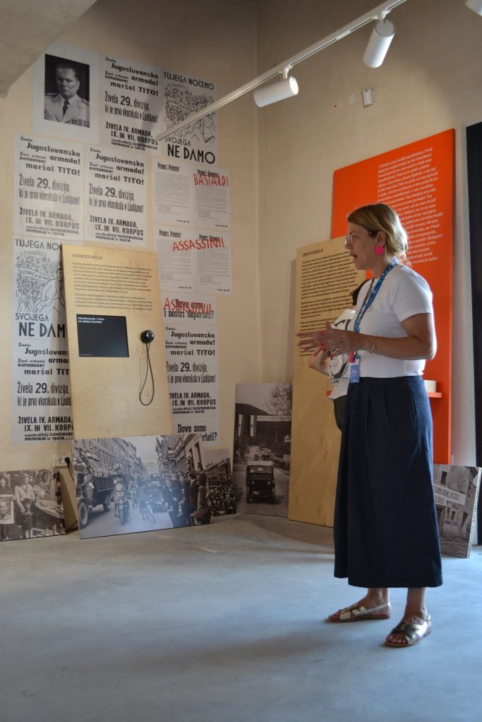 A woman stands in a museum exhibit with historical posters, photos, and text on the walls. She holds papers and wears a white shirt, dark skirt, sandals, and pink earrings. A phone is mounted on a board behind her.