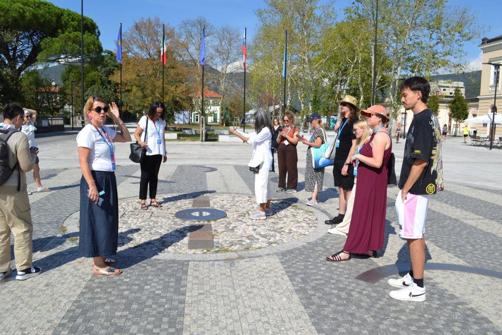 A group of people stands in a circle outdoors on a patterned stone plaza, listening to a guide who is speaking and gesturing. Trees, flags, and buildings are visible in the background.