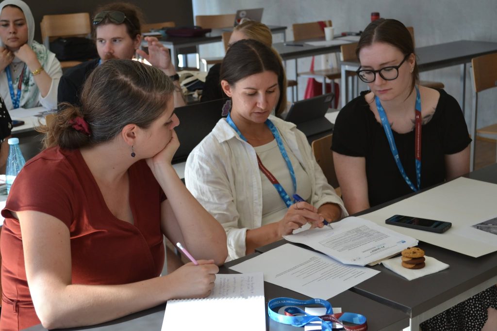 Three women sit at a table, collaborating on documents. They are in a classroom with other people, laptops, and papers visible. Sunlight streams in, and a few snacks are on the table.