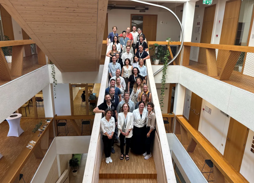 A group of about 30 people stands posing for a photo on a wooden staircase inside a modern building with white walls, wooden floors, and glass doors. Plants hang from the upper floors.