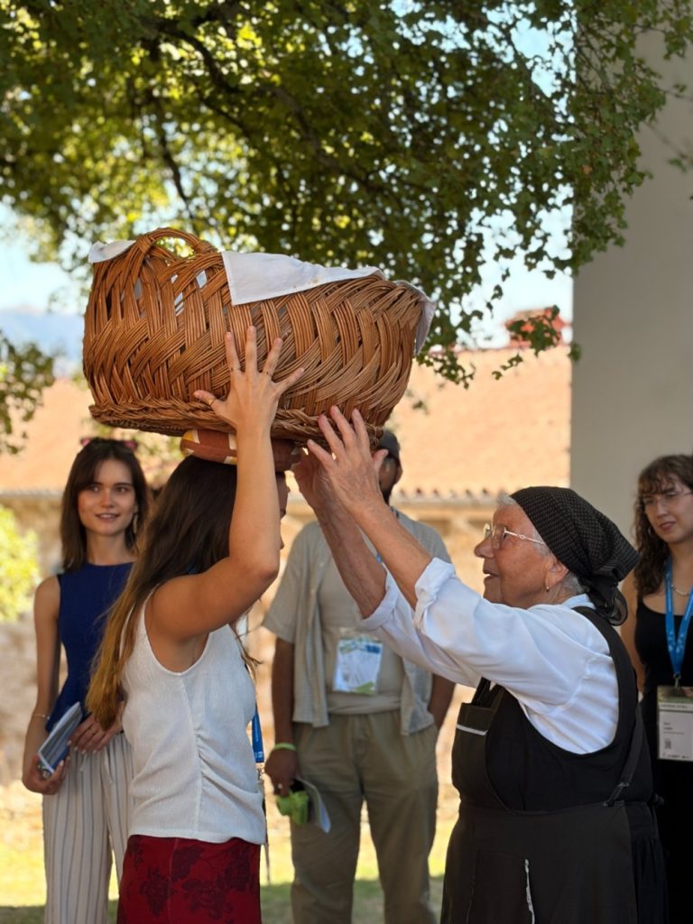 An elderly woman helps a younger woman balance a large woven basket on her head outdoors, while others watch in the background.