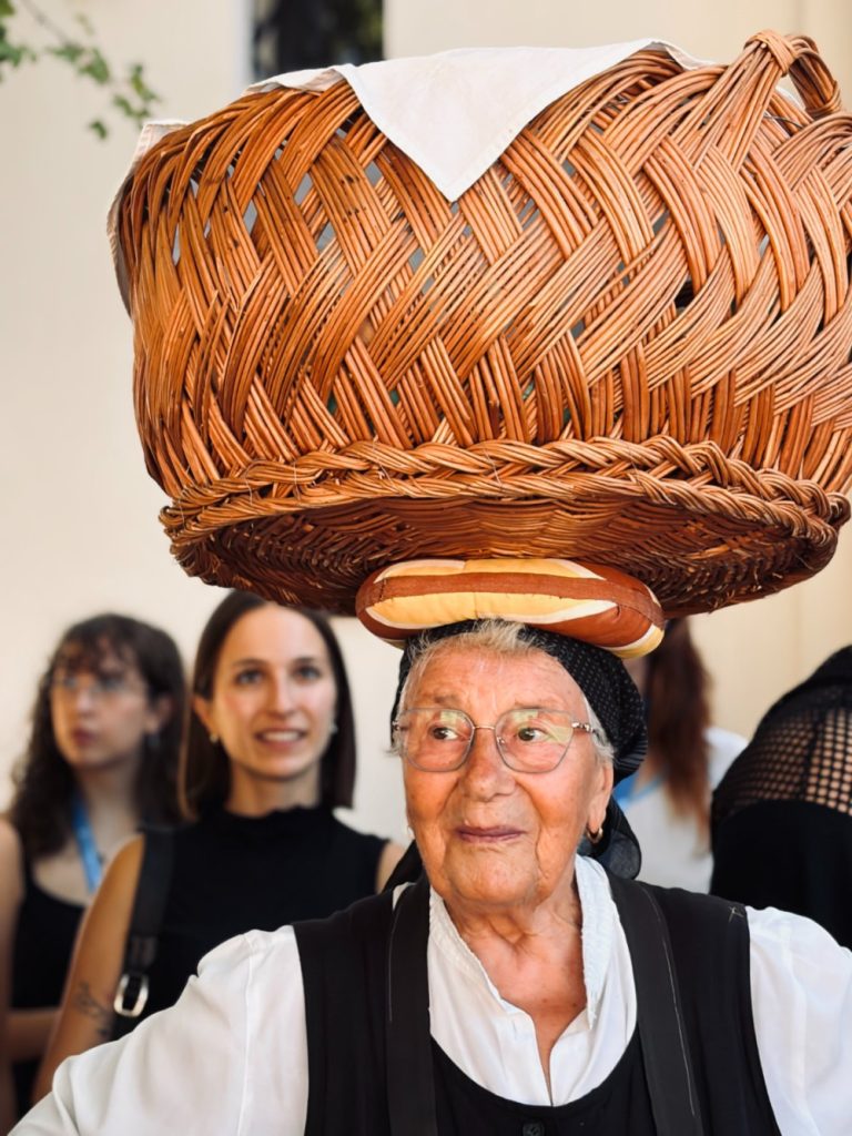 An elderly woman wearing glasses balances a large woven basket on her head, while several people stand in the blurred background.
