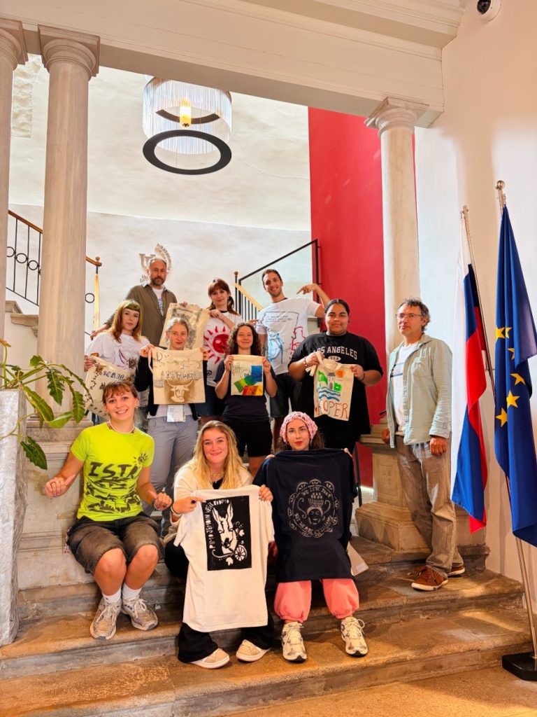 A group of people pose on stairs indoors, holding up decorated shirts and artwork. Two flags stand on the right, and there are marble columns and a red accent wall in the background.