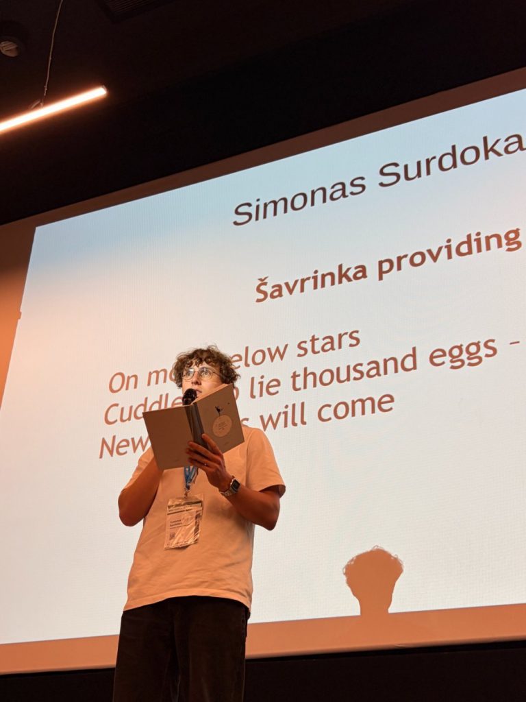 A person stands on stage holding a microphone and booklet, reading in front of a screen displaying poetry and the name Simonas Surdoka. The persons shadow is visible on the screen.