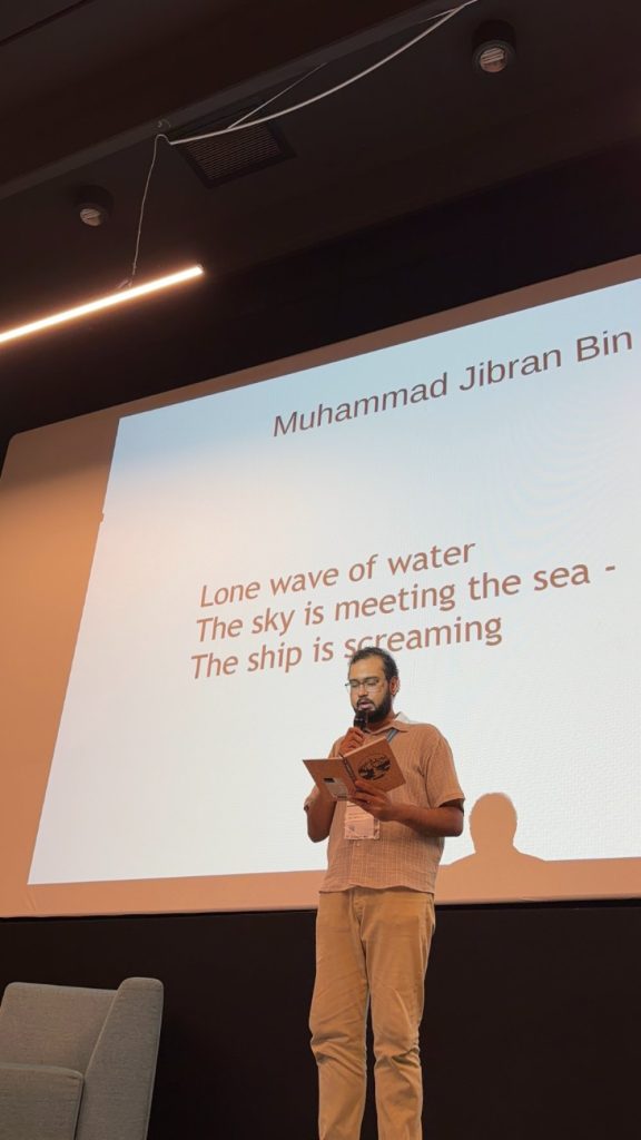 A man stands on stage reading from a tablet, with a poem and the name Muhammad Jibran Bin projected behind him. The poem reads: Lone wave of water / The sky is meeting the sea - / The ship is screaming..