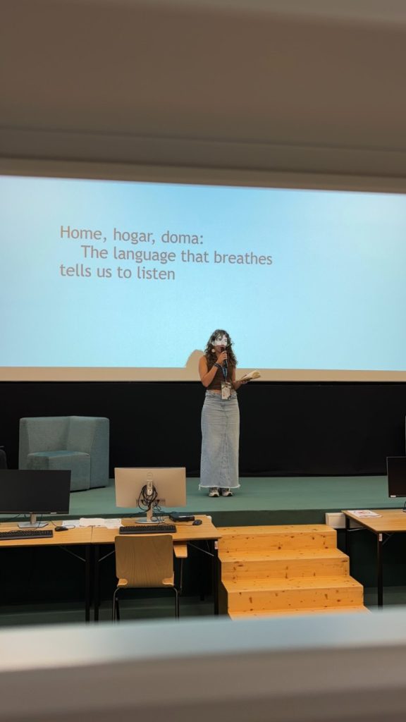 A woman stands on stage speaking, with a large screen behind her displaying text about language and listening. There are chairs, computers, and wooden steps in the foreground.