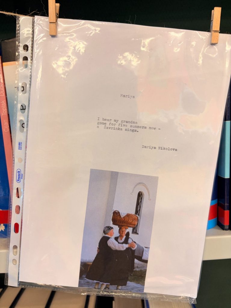 A printed sheet with a short poem typed on it, clipped to a line. Below the poem is a color photo of two women by a white building, one carrying a basket on her back. Books are visible in the background.