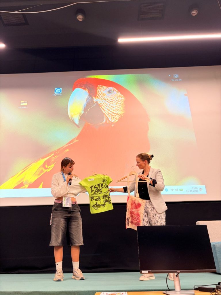 Two people stand on stage in front of a large screen displaying a parrot, holding up a bright green T-shirt and a tote bag, appearing to present or exchange the items during an event or presentation.