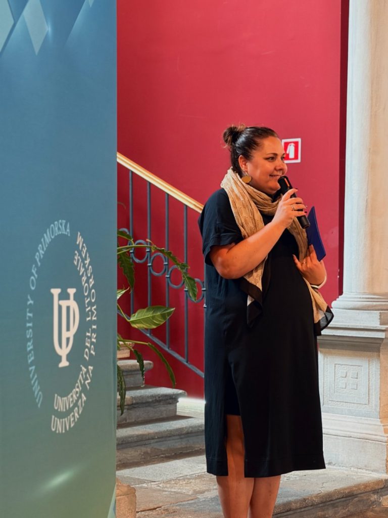 A woman in a black dress and tan scarf speaks into a microphone, standing beside a University of Primorska banner. She is on stone steps with a red wall and a potted plant in the background.
