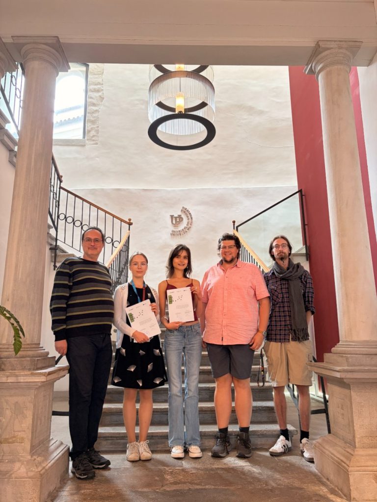Five people stand on stairs in a bright, elegant hall with columns and a chandelier. Three people in the center hold certificates, while two others stand on either side. All are facing the camera and smiling.