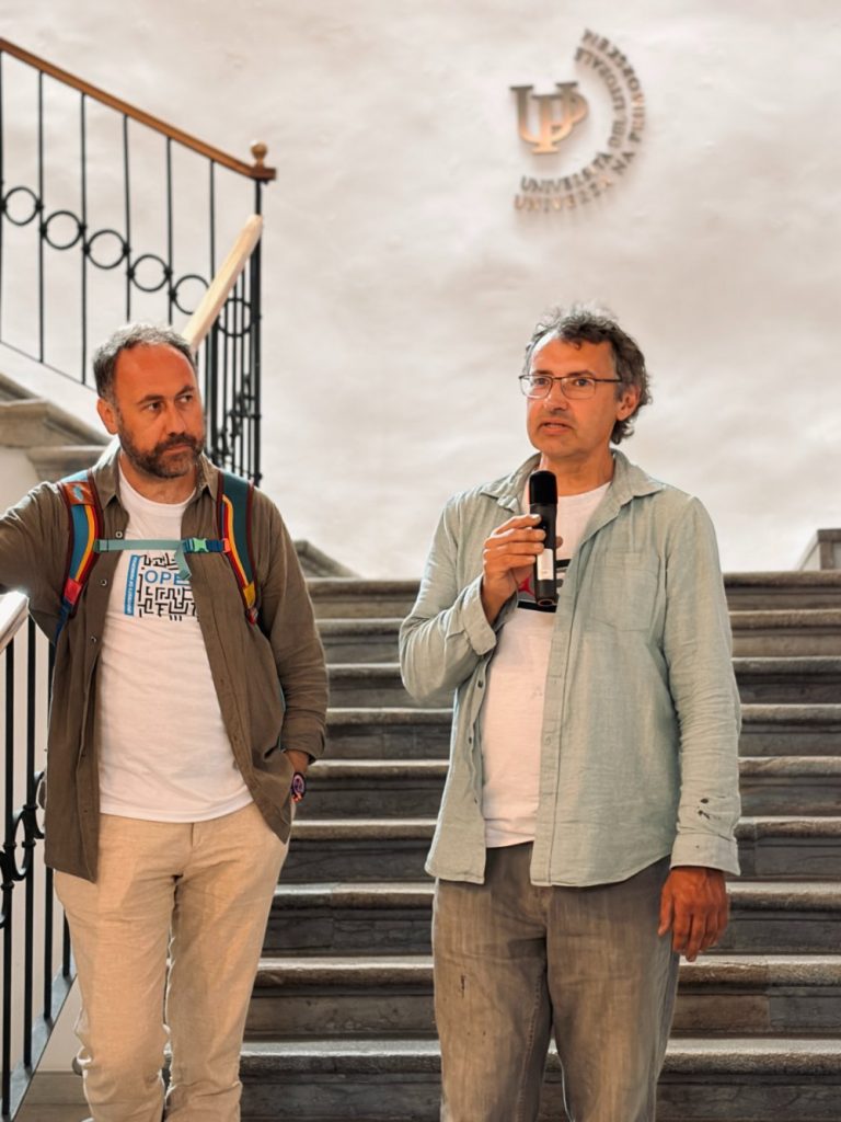 Two men stand on stairs; one holds a microphone and speaks while the other listens. Both wear casual clothes. A university logo is visible on the white wall behind them.