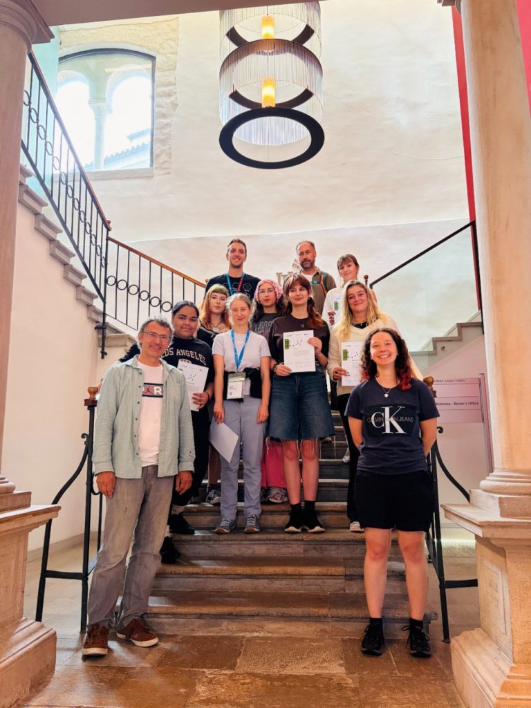 A group of people pose on a staircase inside a bright building, some holding certificates. A round ceiling light hangs above them and sunlight streams through a window on the left.