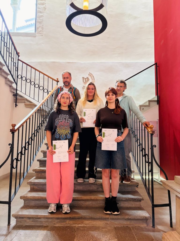 Five people stand on a staircase indoors. Three young women in the front hold certificates, while two adults stand behind them. The setting includes red and white walls and a decorative light fixture.