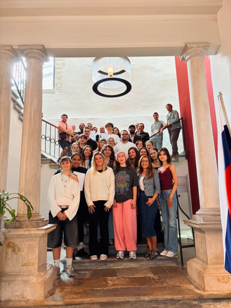 A group of people pose for a photo on a staircase in a bright building with columns and a hanging circular light fixture. Some stand at the bottom while others are on the stairs. A flag is visible to the right.
