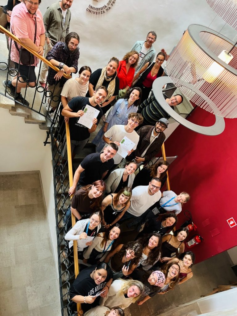 A group of people pose on a staircase, some holding certificates. They are smiling and looking up at the camera. A chandelier hangs above, and a red wall is in the background.