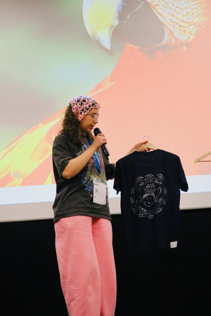 A person in pink pants and a headscarf speaks into a microphone and holds up a navy blue T-shirt on a hanger, standing in front of a large projected image of a parrot.