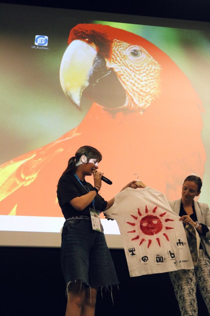 Two women stand on stage; one speaks into a microphone while holding a shirt with a red sun and symbols. The other woman holds the shirt. A large image of a colorful parrot is displayed on the screen behind them.