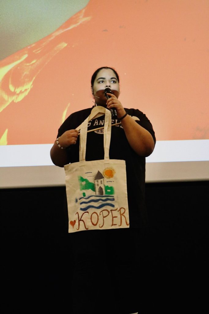 A person stands on stage holding a microphone and a white tote bag decorated with drawings and the word KOPER. The background features a red and white projection screen.