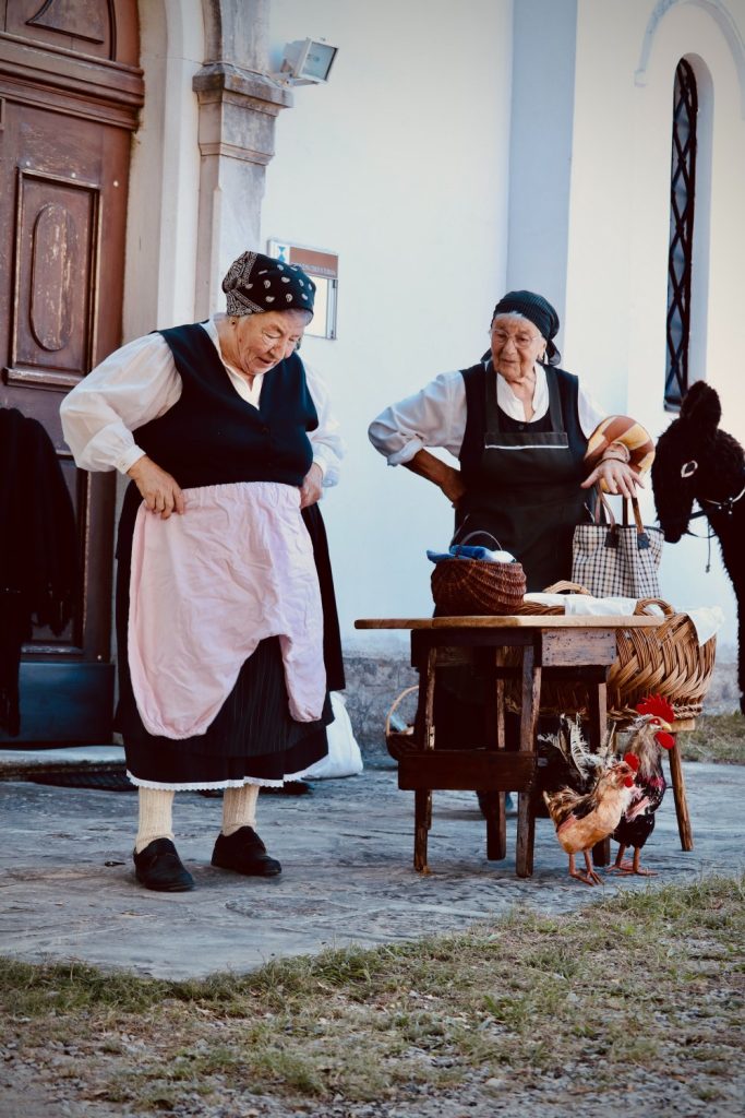 Two elderly women in traditional clothing stand by a wooden table with baskets and a black donkey nearby. Roosters are on the ground, and the scene appears to be outside a rustic building.