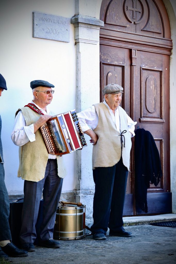 Two elderly men in traditional clothing stand outside a building; one plays an accordion while the other stands with a hand on his hip. Both wear flat caps and vests, with a drum on the ground nearby.