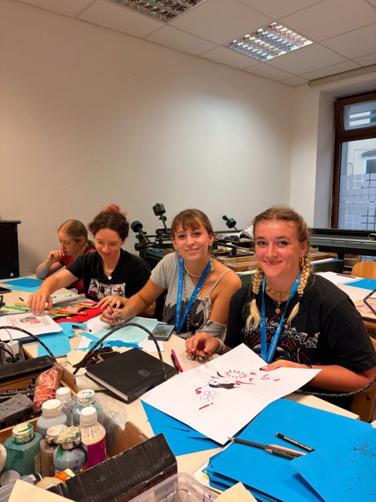 Four young people sit at a table, drawing and painting on paper. Art supplies, sketches, and blue folders are spread around. They smile at the camera in a bright classroom with windows and shelves in the background.