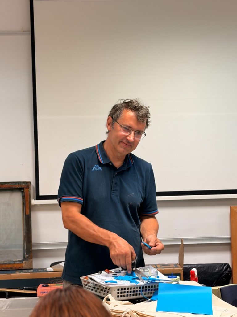 A man in glasses and a navy polo shirt stands at a table sorting items in a basket, with a whiteboard and various supplies behind him.