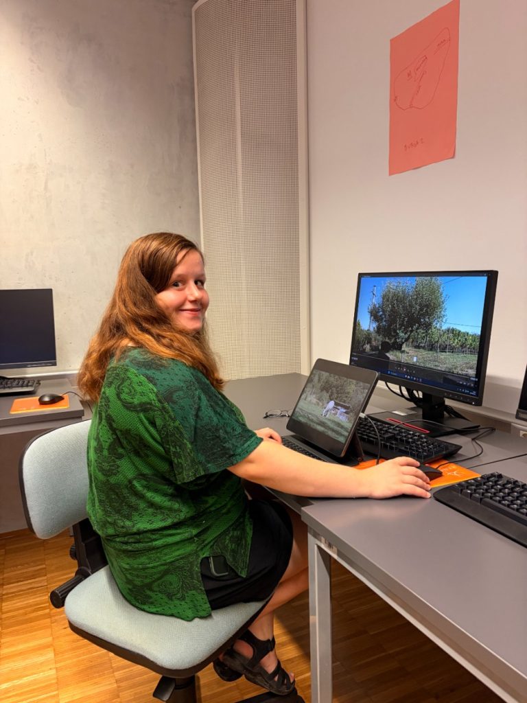 A person with long brown hair, wearing a green shirt, sits at a desk using a laptop and a desktop computer, both displaying outdoor scenes. The person looks back and smiles at the camera.