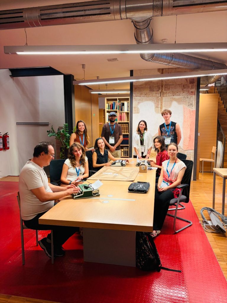Nine people are gathered around a conference table with laptops and notebooks in a modern office space, featuring shelves, stairs, and red flooring. Some are seated, others are standing, all facing the camera and smiling.