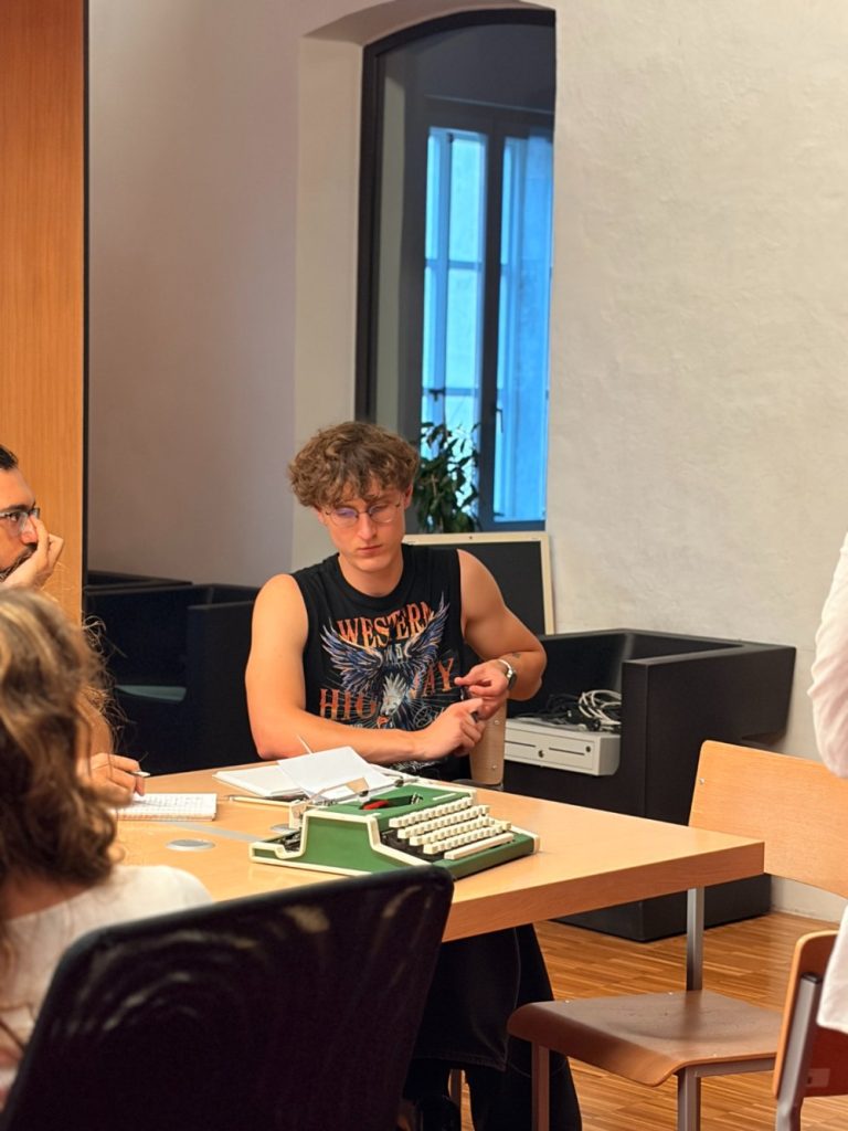 A person with curly hair and glasses sits at a table with a typewriter, wearing a sleeveless shirt. Others are seated nearby, and light enters through a window in the background.