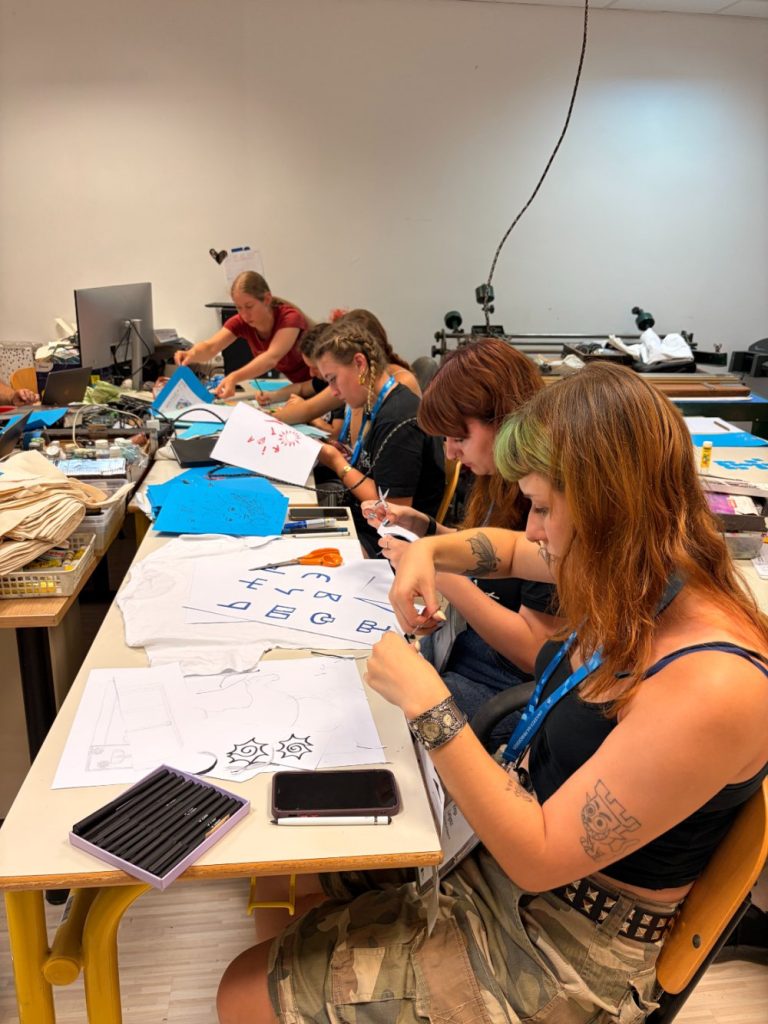 Four people sit at desks working on art projects with paper, pens, and other materials in a classroom setting. Art supplies and tools are scattered on the tables. A computer and various equipment are visible in the background.