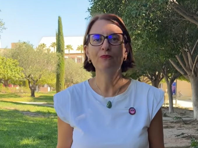A person with short brown hair and glasses stands outdoors in a park, wearing a white shirt and a green pendant necklace. Trees and a building are visible in the background on a sunny day.