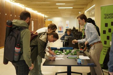 People stand and sign papers at a registration table indoors. One person hands out lanyards and badges. Other attendees wait in line. Office supplies and forms are on the table. A green conference banner is visible.