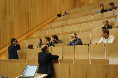 People sit and talk in a mostly empty lecture hall with wooden seats. Some use laptops, while others converse in small groups. A presenter stands near the front, facing the attendees.
