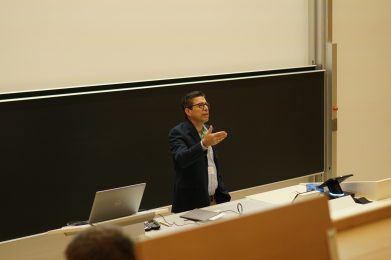 A man in a suit gestures while speaking at a podium in a lecture hall. A laptop, papers, and microphone are on the desk in front of him, with a large blackboard behind.