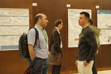 Four people wearing conference badges stand and talk in front of scientific research posters on a brown wall. Two men in the foreground are engaged in conversation, both carrying bags. The background is slightly out of focus.