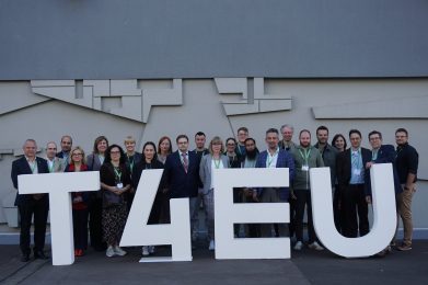 A group of people stand behind large white letters spelling T4EU outdoors, smiling at the camera. Most wear green lanyards, and a geometric gray wall is visible in the background.