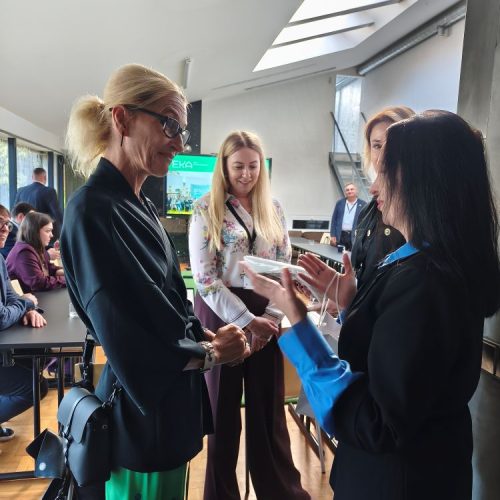 Four women stand and talk indoors at a table, one holding a phone. Other people are seated in the background. The space is bright with large windows and natural light.
