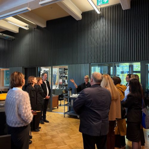 A group of people stand in an office space while one person gestures, appearing to give a tour or presentation. The room has wooden floors, black walls, and large windows in the background.