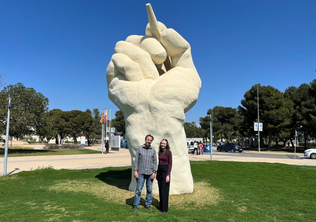 Two people stand smiling in front of a large white sculpture of a raised fist holding a pencil, outdoors on a sunny day with trees and flags in the background.