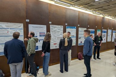 People stand in front of scientific posters displayed on a wall, engaging in discussions and presentations at an academic conference or research event.