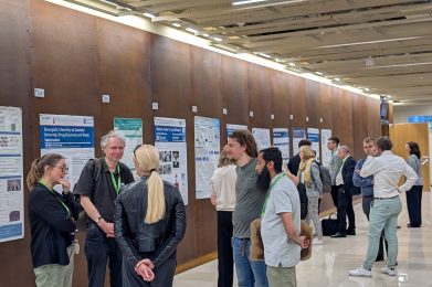 People converse and examine scientific posters displayed on a brown wall in a brightly lit hallway during a conference or academic event.