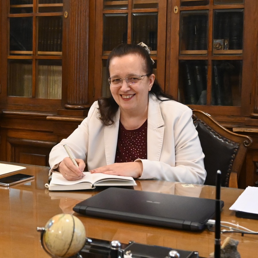 A woman in a light blazer sits at a wooden desk, smiling while writing in a notebook. Behind her are bookshelves with glass doors filled with books. A globe and a closed laptop are on the desk.