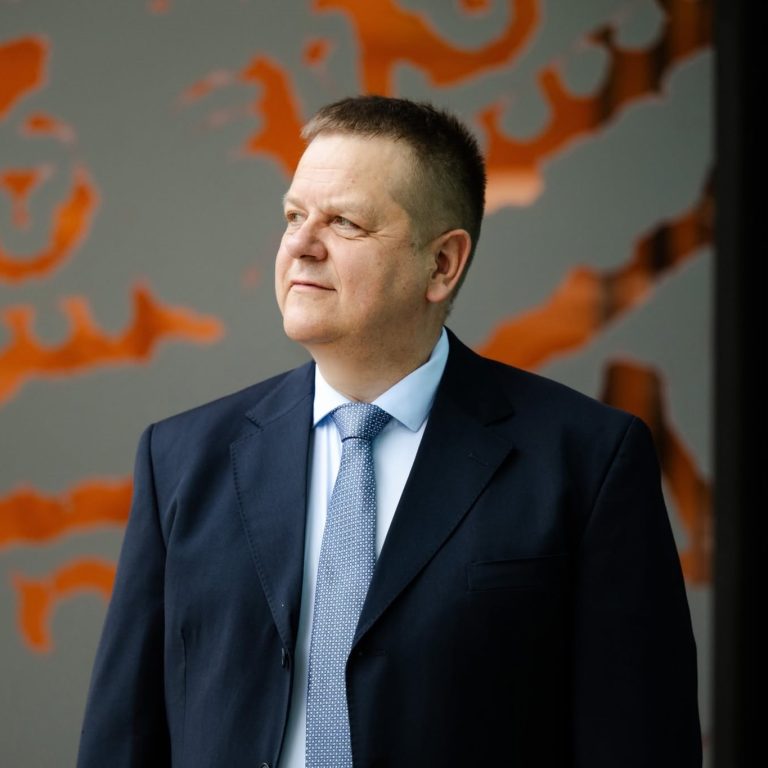 A man in a navy suit and light blue tie looks to his left, embodying a poised confidence. The background features a gray wall with abstract orange patterns, evoking the dynamic spirit to transform for Europe.