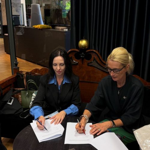 Two women sitting at a small round table, signing documents together in a modern indoor setting with dark curtains and various office furniture in the background.