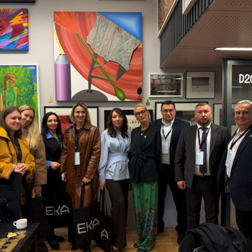 A group of ten people pose indoors in front of colorful artwork, wearing business or smart casual attire with conference lanyards. Some hold black bags labeled EKA. A door labeled D207.2 is visible to the right.