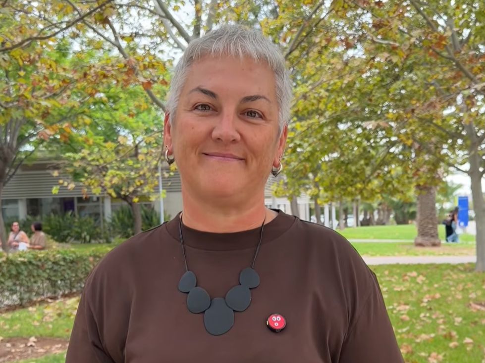 A person with short gray hair, wearing a brown shirt, black necklace, and a red pin, stands outdoors in a park-like area with green trees and a building in the background.