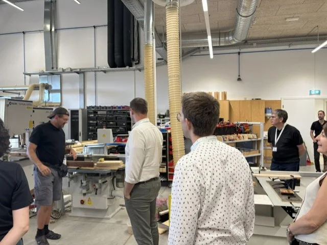 A group of people stand in a workshop with woodworking machines, observing a man explaining something. Tools and materials are visible on shelves and benches in the background.